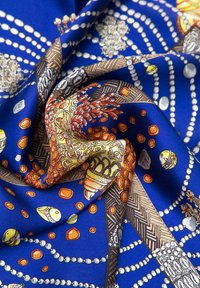 Tessuto di seta blu con motivi intricati, tra cui conchiglie, perline e forme astratte in arancione, giallo e bianco. Texture liscia.