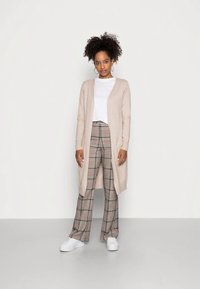 Sieviete ar cirtainiem matiem, ģērbusies beignet garajā kardiganā, baltā krekliņā, raibajās platās biksēs un baltos sporta apavos, stāv uz vienkārša balta fona.