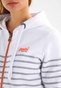 Haut à zip blanc avec des rayures grises et un logo orange. Dispose d'une capuche avec cordon de serrage et d'une fermeture éclair métallique. Le tissu semble doux et texturé.