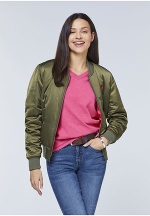 Olivegrüner Bomberjacke mit glänzender Textur, Reißverschluss und gerippten Bündchen. Kombiniert mit einem pinken V-Ausschnitt-Pullover und blauen Jeans.