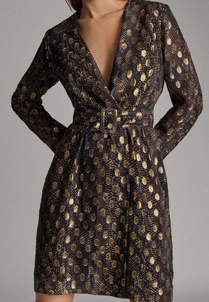 Femme portant une robe portefeuille noire à pois dorés avec une ceinture assortie, debout les mains dans le dos.