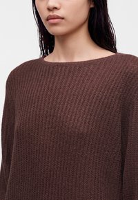 Jeune femme aux cheveux lisses et foncés portant un pull en maille marron texturé avec un large col rond sur un fond uni.