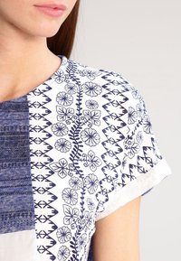 Blus med en blandning av blå och vita tyger, blommiga och geometriska mönster, korta ärmar, rund hals och en texturerad design.