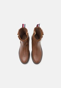 Botas de cuero marrón hasta el tobillo con un acabado suave y punta redondeada. Presentan suelas negras y una lengüeta a rayas en la parte trasera. Acentos en dorado.