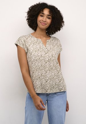 Kvinde med krøllet hår iført en kortærmet mønstret bluse og lyseblå jeans, stående mod en ensfarvet lys baggrund.