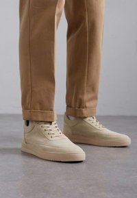 Beige velúr sportcipők alacsony fazonban, fűzős záródással és sima gumitalppal; barnára felhajtott chinos nadrággal párosítva.