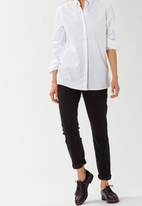 Camisa blanca de botones con mangas enrolladas, combinada con pantalones negros con dobladillo y zapatos oxford negros con detalles de tachuelas.