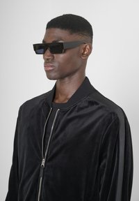Chaqueta bomber negra de terciopelo, con cremallera plateada, cuello acanalado y rayas negras en las mangas. Gafas de sol cuadradas negras.