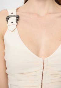 Bežinis be rankovių palaidinė su giliu V formos iškirpimu, su raukšlėtu audiniu ir sidabriniu apdailos elementu, kuriame užrašyta "Versace Jeans Couture" ant petnešėlės.