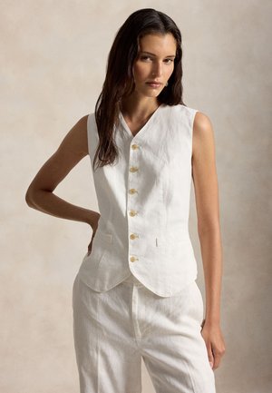 Nő ujjatlan fehér, gombos mellényben és hozzá illő fehér nadrágban, egy kézzel a csípőjén áll semleges háttér előtt.