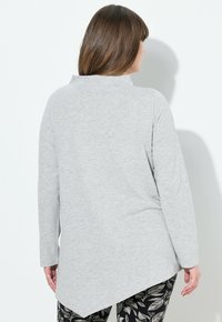 Graues Langarmoberteil mit hohem Kragen und asymmetrischem Saum. Weicher Stoff. Kombiniert mit gemusterten schwarz-weißen Hosen.