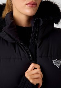 Juodas pūkinis švarkas su dirbtinio kailio kapišonu, su prekės ženklo užtrauktuku ir trikampiu logotipo uždėjimu kairėje krūtinės pusėje. Matomi tekstūruoti megztiniai rankogaliai.