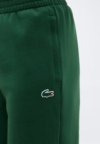 Σκούρο πράσινο φόρμας με ελαστική μέση, πλαϊνή τσέπη και ένα μικρό κεντημένο λογότυπο κροκόδειλου κοντά στην τσέπη.