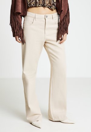 Jean flare beige avec une texture lisse, doté d'une ceinture standard, de poches avant et d'une fermeture par bouton. Associé à une veste marron à franges.