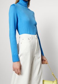 Haut à col roulé bleu en tissu léger, associé à un pantalon blanc à jambes larges et taille haute. Main tenant un sac à main jaune.
