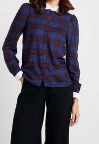 Button-up shirt featuring a blue and burgundy plaid pattern, long sleeves, slight puff shoulders, and a white collared neckline.