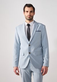 Traje azul claro con un diseño estructurado, dos bolsillos frontales y botones marrones. Combinado con una camisa blanca y una corbata negra. Textura suave.