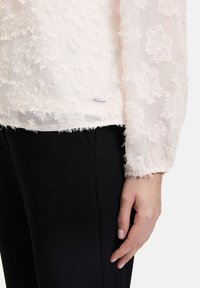 Helle rosa transparente Bluse mit strukturierten, ausgefransten Blumenmustern, elastischen langen Ärmeln, kombiniert mit schwarzen schmalen Hosen. Markenlabel sichtbar.