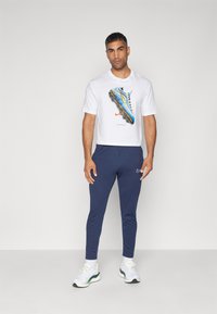 T-shirt grafica bianca con stampa di scarpe da atletica blu, abbinata a jogger affusolati blu navy e sneakers bianche con dettagli verdi.