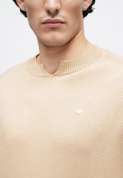 Gros plan sur une personne portant un pull en maille beige avec un petit logo sur la poitrine et de petites boucles d'oreilles clous.