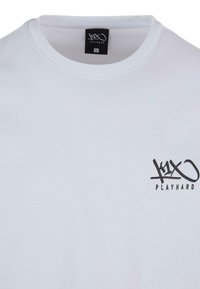 White cotton T-shirt with a round neckline. Features a black printed logo "k1x PLAYHARD" on the left chest area. Size tag "L" visible.