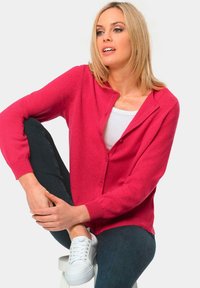 Felle roze cardigan met een knoopsluiting, geribbelde manchetten en een relaxte pasvorm, gecombineerd met een witte top en donkere jeans, gedragen met witte sneakers.