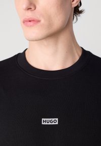 Černá pletená mikina s crew neck a bílým logem "HUGO" umístěným na levé části hrudi. Měkká bavlněná textura.