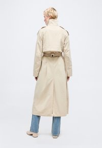 Άτομο με κοντά ξανθά μαλλιά που φοράει μπεζ καμπαρντίνα με ζώνη, μπλε τζιν και μπεζ παπούτσια, στέκεται γυρισμένο με την πλάτη προς λευκό φόντο.