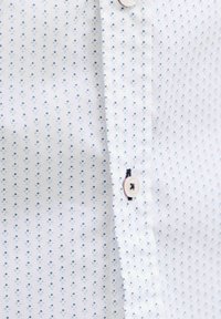 White shirt with small blue diamond pattern, close-up showing white button sewn with dark blue thread.