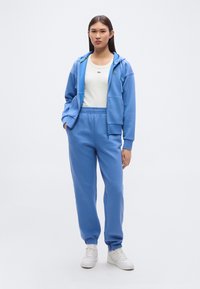 Φωτεινό γαλάζιο φούτερ με κουκούλα και ταιριαστό παντελόνι φόρμας, και τα δύο από βαμβάκι με απαλό υλικό. Λευκό T-shirt από κάτω. Λευκά αθλητικά παπούτσια.