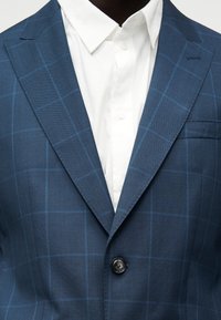 Μπλε blazer με καρό μοτίβο παραθύρου, φορεμένο πάνω από λευκό πουκάμισο με κουμπιά και κλασικό γιακά.