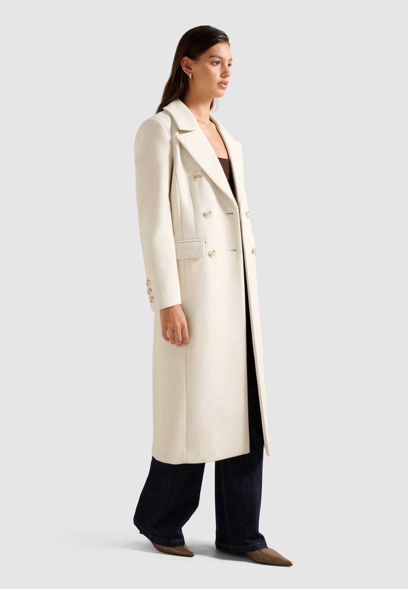 Lungo cappotto bianco doppiopetto con ampi revers e bottoni dorati; abbinato a jeans neri a gamba larga e scarpe beige con punta.