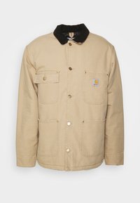 Beige canvasjakke med en sort kordfløjskrave, der har fire frontlommer og guldknapper. Logo-patch på brystlommen.