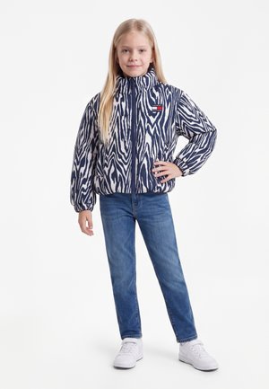 Pige med langt blondt hår iført en blå og hvid mønstret dunjakke, blå jeans og hvide sneakers, står med den ene hånd på hoften.