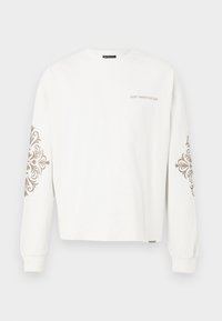 Witte lange mouwen t-shirt van katoen met decoratieve bruine bloemenpatronen op de mouwen en de tekst "VERSPEEL CULTUUR NIET" op de voorkant.