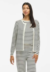 Cardigan a righe bianche e nere con maniche lunghe e apertura con bottoni; abbinato a pantaloni a righe, caratterizzati da un tessuto morbido e testurizzato.