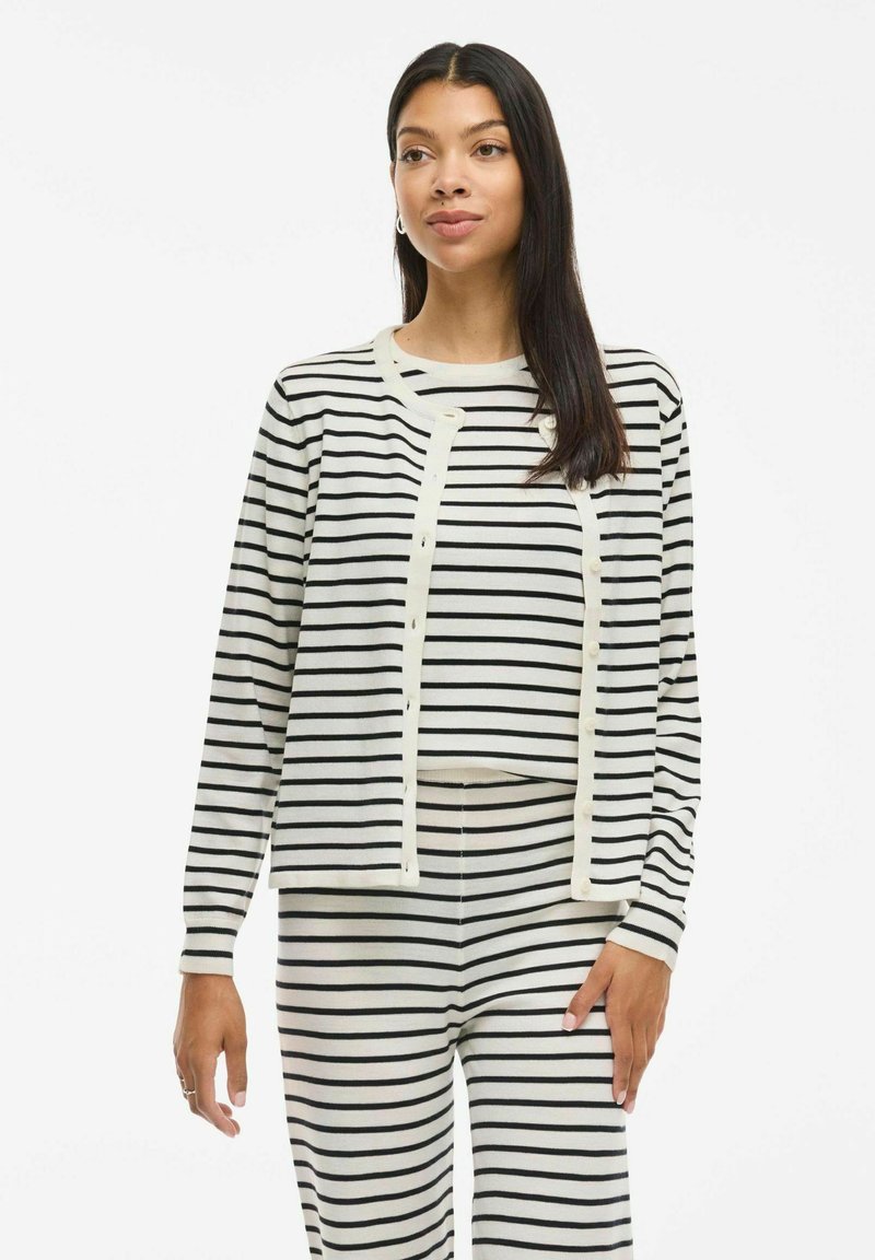 Cardigan a righe bianche e nere con maniche lunghe e apertura con bottoni; abbinato a pantaloni a righe, caratterizzati da un tessuto morbido e testurizzato.