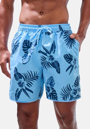 Modré plavkové šortky s tmavomodrými tropickými listami a kvetinovými vzormi, elastickým pásom s bielym sťahovacím šnúrkom, nosené stojacou osobou.