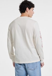 Long-sleeve cream shirt with a round neck, featuring red text along the left sleeve, paired with light blue distressed jeans.