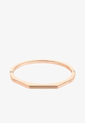 Vaijeri zelta krāsas aproce ar gludu, apaļu formu un plakaniem malām. Virsma ar teksturētiem akcentiem vienā pusē un iegravētu logotipa detaļu.