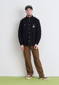 Schwarzes Hemd mit weißen Druckknöpfen, braune Cargo-Hose und beiges Sneakers. Grüner Hut auf dem Kopf, stehend vor einem schlichten Hintergrund.