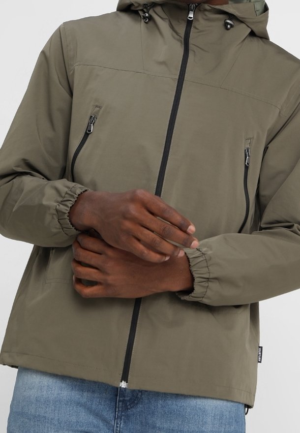 Person i en olivgrön jacka med huva, svart dragkedja och fickor på bröstet, som justerar ärm cuffen.