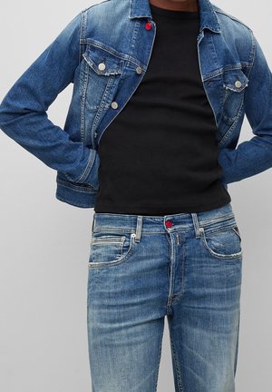 Persoon die een blauwe spijkerjacks, een zwarte geribbelde trui en blauw verweerde jeans met rode knoopdetails draagt, met de handen in de zakken staand.