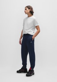 Weißes Baumwoll-T-Shirt mit Rundhalsausschnitt; marineblaue Sweatpants mit elastischem Bund und Seiten Taschen; schwarze Sportschuhe mit roten Akzenten.