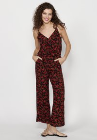 Zwarte bloemen jumpsuit met rode bloemen, v-hals, dunne bandjes en losse wijde benen met zakken. Gecombineerd met platte sandalen.