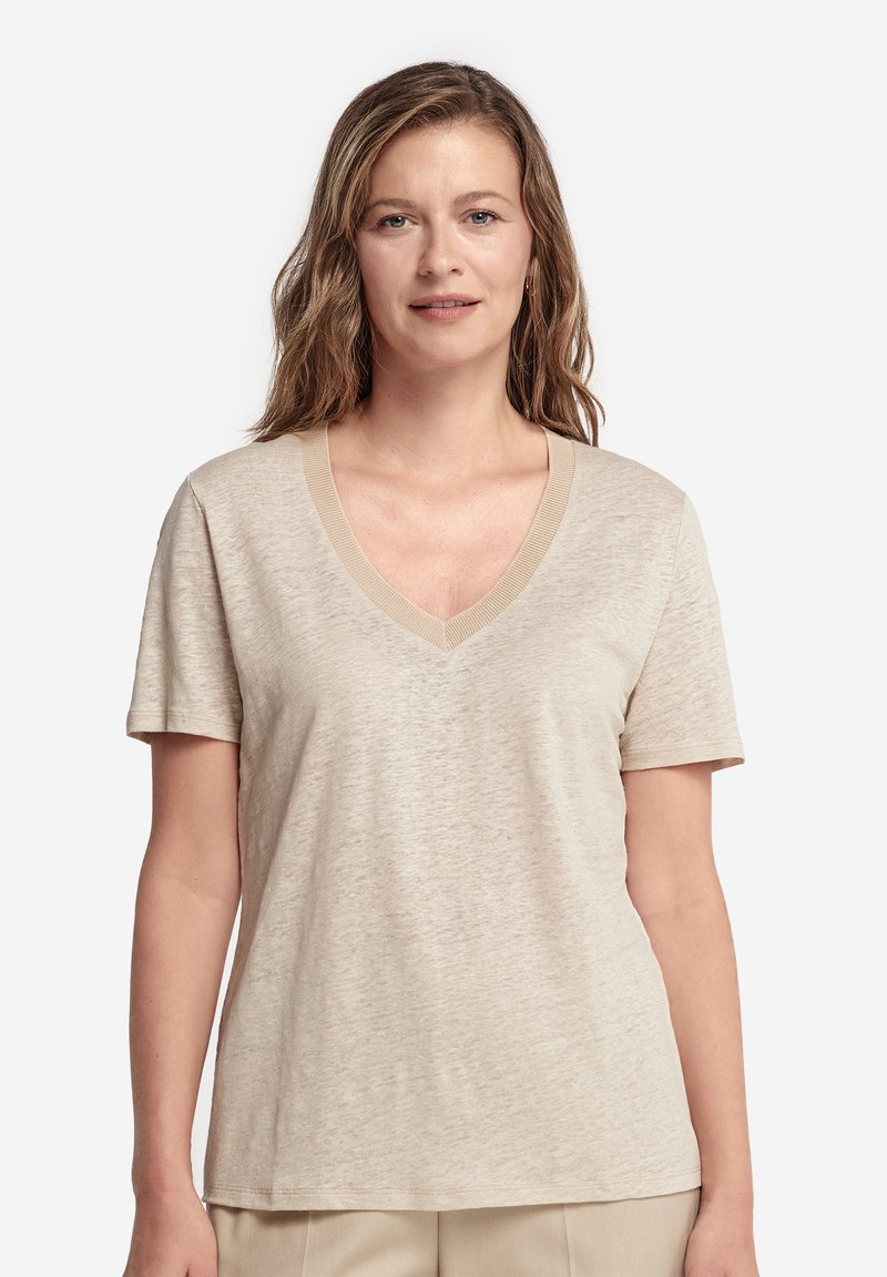 Femme aux cheveux ondulés châtain clair portant un t-shirt beige à manches courtes et col en V, debout devant un fond clair uni.