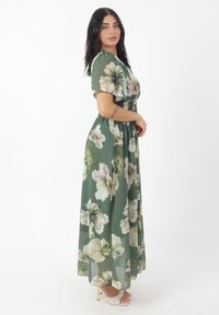 Robe maxi florale verte avec des manches courtes, taille smockée et tissu fluide. Présente de grandes fleurs blanches et vert clair. Texture légère.