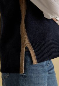 Granatowy sweter z kontrastowym beżowym panelem bocznym, z prążkowanym wykończeniem i rozcięciem. Noszony na jasnoniebieskich dżinsach, podkreślający teksturę i warstwy.