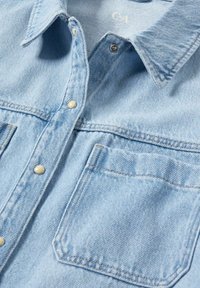 Chemise en denim bleu clair avec boutons-pression, une poche poitrine et un col, étalée à plat montrant la texture du tissu et les détails des coutures.