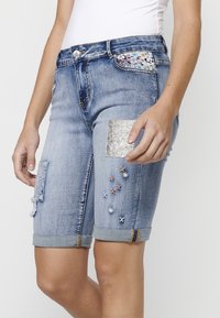 Shorts en denim de couleur bleu clair avec des ourlets retroussés, décorés de divers patchs, d'accents en sequins et de détails brodés colorés sur le devant.